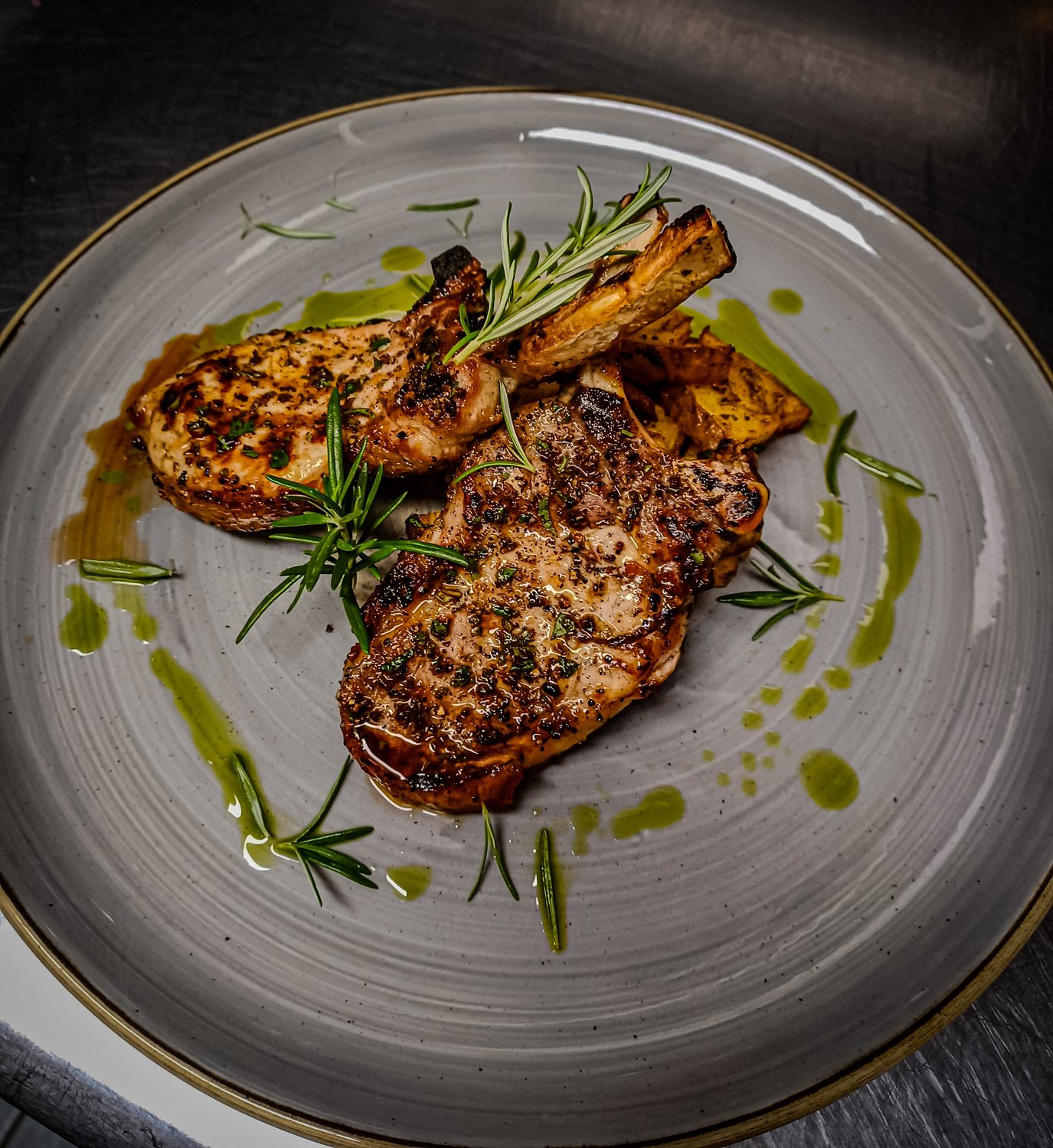 Grilled pork tomahawk steaks on the bone with rosemary, roast potatoes and a drizzle of herb oil on a stoneware plate