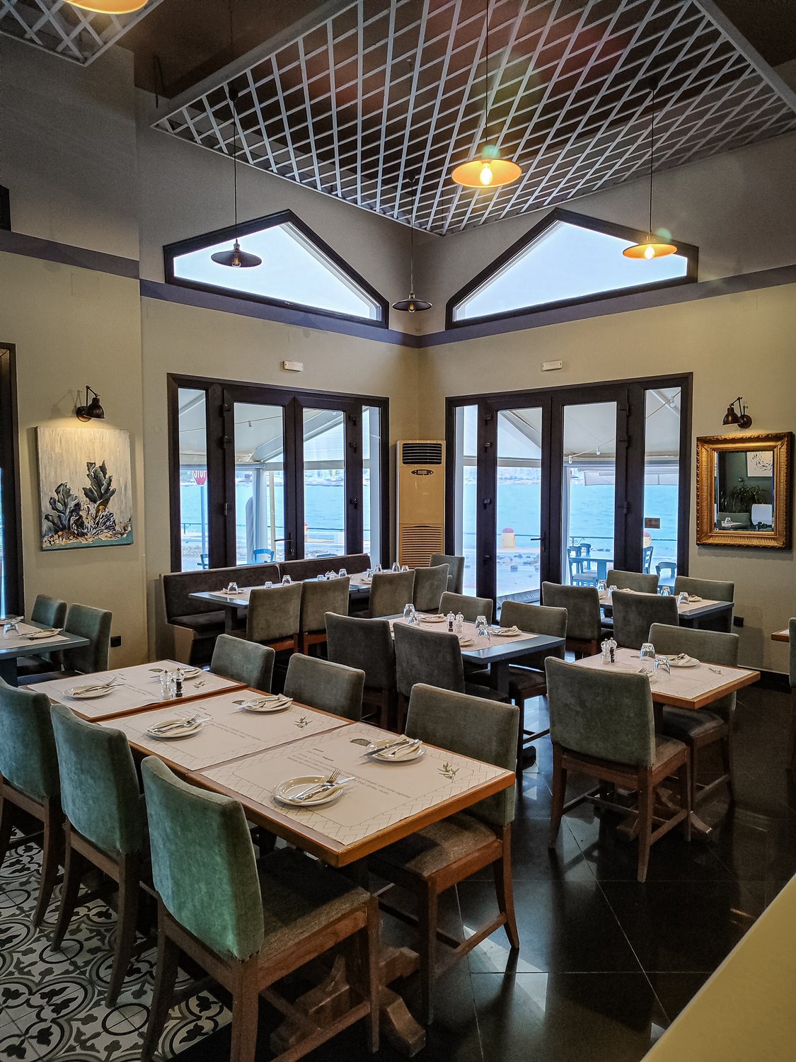 The dining room inside El Greco Leros, looking through glass doors that open onto the waterfront terrace and the sea