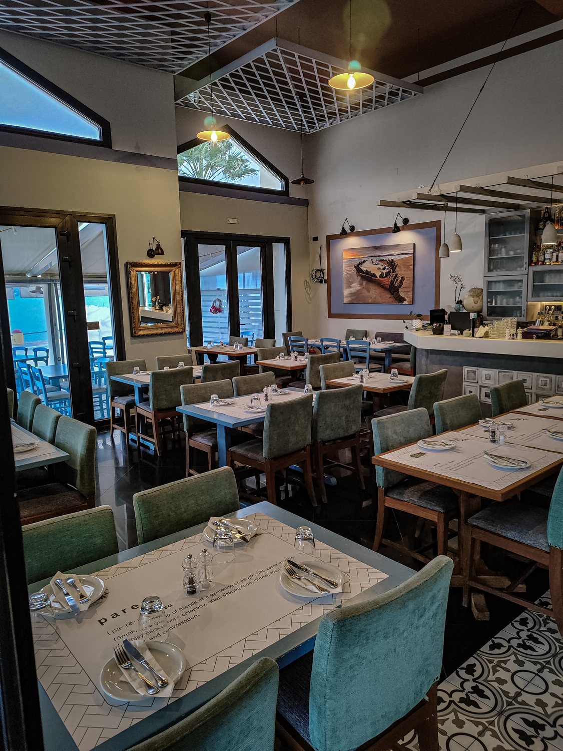The dining room with green upholstered chairs, blue tables, patterned floor tiles and the bar at the far end