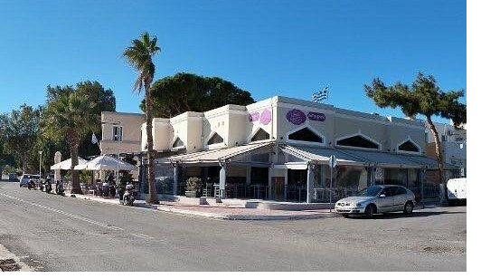 The whitewashed exterior of El Greco Leros with palm trees, awnings and shaded pavement tables under a clear sky